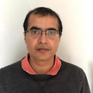 Dr. Mishra sports a pink collar over a grey sweater. Close cropped hair and bespectacled, Dr. Mishra stares into the camera with a contemplative expression. 