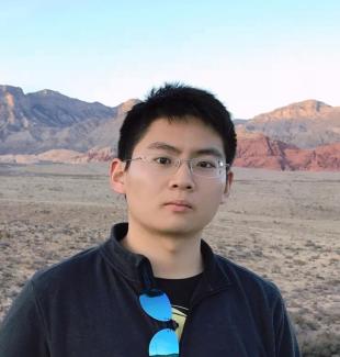 Dr. Gengchen Mai stands in from of a rock expanse wearing a black jacket, glasses with metal frames, and sunglasses on his jacket collar. He stares into the camera without smiling. 