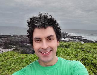 Dr. Santiago Herrera, backed by a rocky coast line, smiles with salt and pepper curly hair in a bright green shirt. 