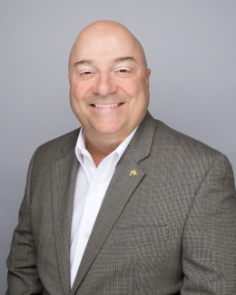 Dr. Sutton smiles widely to the camera. He is wearing a grey suit jacket, white button up, and a golden shark pin. 