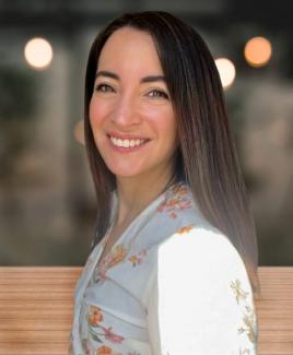 Dr. Valerie De Anda stands sideways with her head turned towards the camera smiling. Her dark brown hair falls behind her back against her white button up. 