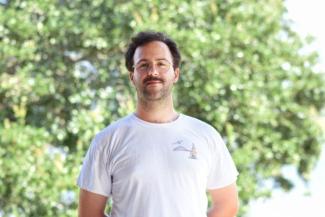 Ben Lowin stands in a white t-shirt in front of green foliage. Fluffy dark hair halos his face, speckles his jaw, and condenses in a mustache. He looks proud though his spectacles and into the camera.  