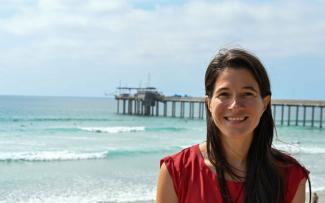 Dr. Moira Decima smiles at the camera in a shoulderless red shirt backed by the ocean and a pier. 