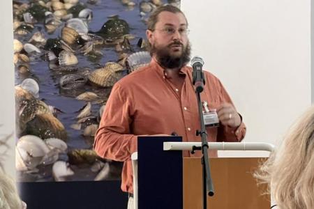 Dr. Philip Riekenberg lectures in a salmon button up in front of a poster of oysters. 