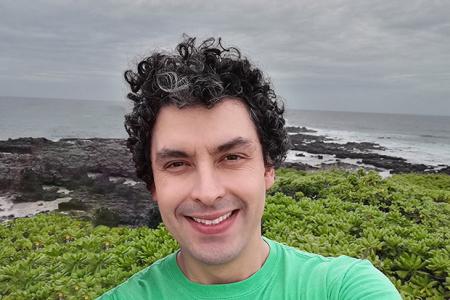 Dr. Santiago Herrera, backed by a rocky coast line, smiles with salt and pepper curly hair in a bright green shirt. 