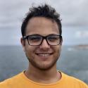 Dr. Coleman smiles at the camera in front of the ocean. He sports a yellow shirt and square black glasses. 