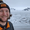 Dr. Zac Cooper appears smiling in the left corner wearing a orange and grey beanie with matching jacket. Behind him is an expanse of frozen water.