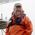 Dr. Sam Bentley smiles at the camera in a orange slicker with emergency life jacket and tan baseball cap