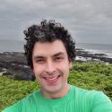 Dr. Santiago Herrera, backed by a rocky coast line, smiles with salt and pepper curly hair in a bright green shirt. 