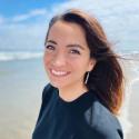 Dr. Valerie De Anda smiles into the camera on the beach with dark red-brown hair blowing in the wind