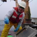 Dr. Tracey Sutton with blue gloves holds a small squid toward the camera. He smiles wearing a white safety helmet and orange safety life vest, with bright yellow pants